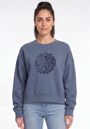 Frau trägt einen blauen Pullover mit einer Grafik von halb Gehirn und halb Blumen und Text "DON'T OVER THINK IT", stehend vor weißem Hintergrund.