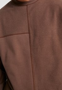 Felpa marrone con due pannelli di tessuto testurizzato uniti da cuciture visibili sul retro, mettendo in evidenza il contrasto dei tessuti e i dettagli delle cuciture.