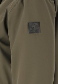 Olijfgroene jas met een gestructureerde stof, elastische manchetten en een zwarte rubberen logo patch met een bergontwerp op de mouw.