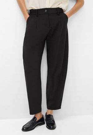 Femme portant un pantalon noir taille haute sur mesure avec des plis à l'avant et des mocassins en cuir noir, mains dans les poches, sur fond blanc.