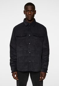 Mörk manchesterjacka med krage, tryckknappar och två bröstfickor. Har en vertikalt ribbad textur och en avslappnad passform.