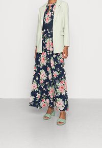 Navy sininen kukkamekko, jossa on vaaleanpunaisia ja vihreitä ruusuja, yhdistettynä vaaleanvihreään bleiseriin. Kudotut mintunvihreät korkokengät täydentävät asun.