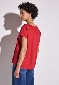 Blusa rossa a maniche corte con collo rotondo e dettaglio a pieghe sul retro, abbinata a jeans in denim blu. Tessuto morbido, vestibilità rilassata.