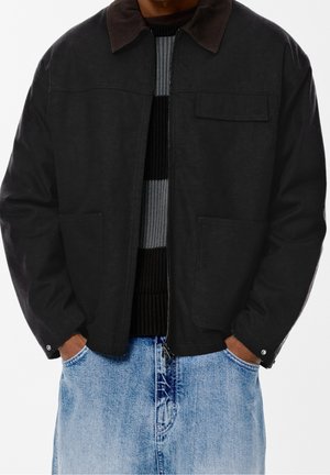 Personne portant une veste noire zippée avec un col sombre sur un pull rayé et un jean en denim bleu clair, les mains dans les poches.