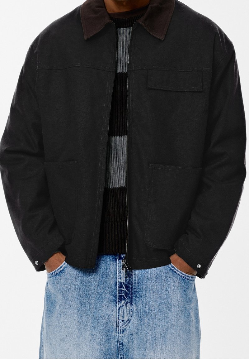 Personne portant une veste noire zippée avec un col sombre sur un pull rayé et un jean en denim bleu clair, les mains dans les poches.