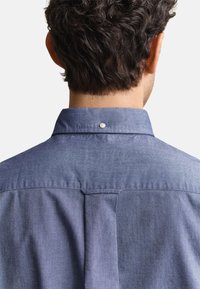 Bărbat cu păr scurt și creț purtând o cămașă albastră cu nasturi, arătat din spate cu un mic nasture alb pe guler.