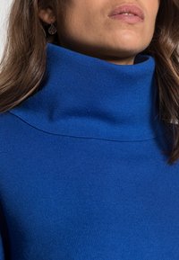 Un pull col roulé bleu à côtes avec un col épais et haut. Le tissu semble doux et texturé, soulignant une coupe décontractée autour du cou.