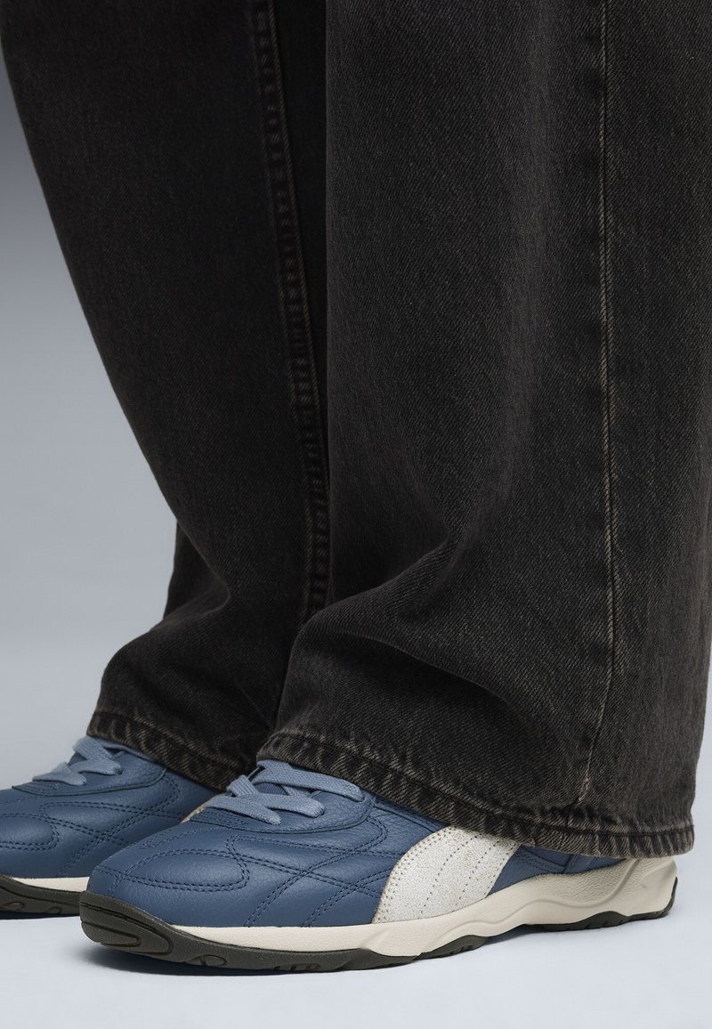 Sapatilhas de pele azul com detalhes brancos e design texturizado, combinadas com jeans de ganga escuros, visíveis nos tornozelos. Fundo preto.