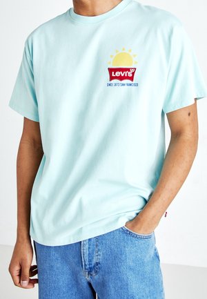 Personne portant un t-shirt Levi's bleu clair avec un motif soleil et un jean denim bleu, main dans la poche sur fond blanc.