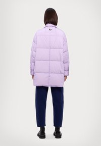 Chaqueta acolchada púrpura con un diseño cuadrado, con costuras horizontales y una pequeña etiqueta en la parte posterior. Lleva puesta con pantalones oscuros azules y botas.