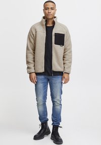 Fleece jacket in beige with a black zippered chest pocket; paired with blue jeans and black lace-up boots. Smooth, textured fabric.