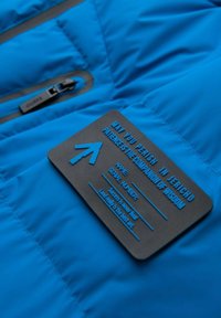 Sininen toppatakki, jossa on sileä, kiiltävä pinta. Siinä on musta merkki, jossa on kohokuvioituja tekstejä, mukaan lukien nuoli ja useita kirjoituksia.