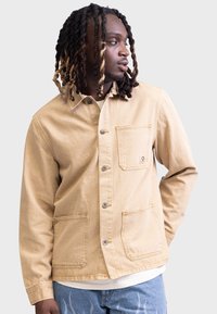 Chaqueta beige con cuello, hecha de algodón, que cuenta con dos bolsillos frontales, cierre de botón y un logotipo sutil en el bolsillo. Tejido ligeramente texturizado.