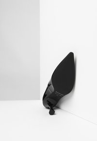 Zapato de tacón alto de charol negro con punta afilada y tacón delgado, que muestra una textura suave y un diseño mínimo en la suela.