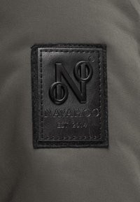 Zwarte leren patch met een reliëf "N" en cirkelontwerp, bevestigd aan een gestructureerde grijze stoffen achtergrond, gelabeld "NAVAHOO EST 2014."