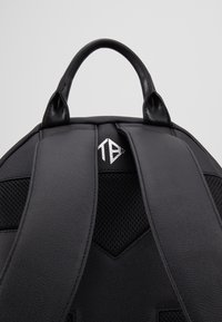 Sac à dos noir avec des bretelles rembourrées, une poignée de transport sur le dessus et un petit logo blanc entre les bretelles.