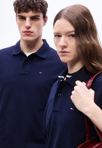 Δύο νέοι ενήλικες φορούν μπλε μπλούζες πόλο με μικρά λογότυπα, ο ένας κρατάει το λουρί μιας κόκκινης τσάντας, και οι δύο κοιτούν σοβαρά μπροστά.