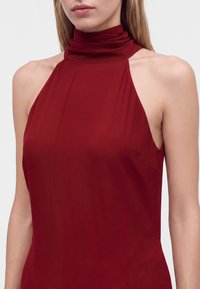 Femme portant une robe bordeaux sans manches à col haut avec col drapé, montrant des épaules et le haut de la poitrine nus sur un fond clair uni.