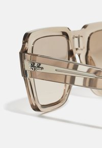 Ochelari de soare Ray-Ban cu un cadru din plastic bej transparent, lentile maro cu nuanță și un detaliu metalic logo pe lateral.