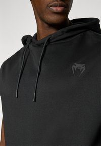 Μαύρο αμάνικο φούτερ με κουκούλα, διαθέτει κορδόνι και διακριτικό λογότυπο φιδιού στο στήθος, κατασκευασμένο από λείο, υφασμάτινο ύφασμα με υφή.