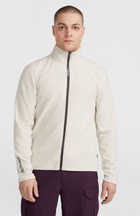 Giacca in fleece crema con colletto alto, zip frontale completa e accessori neri. Presenta il marchio "Polartec" sulla manica.