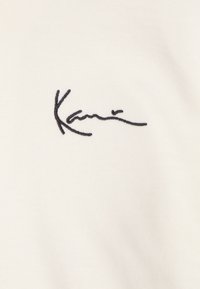 Tejido de algodón blanco con una textura suave, que presenta un diseño de firma bordado en negro sobre un fondo sutil.