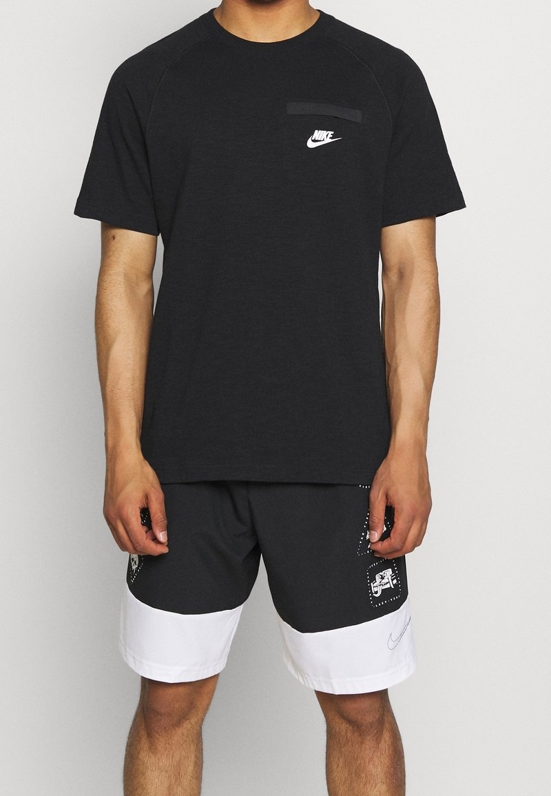 Musta lyhythihainen Nike-t-paita, jossa on valkoinen logo rinnassa, yhdistettynä harmaisiin ja valkoisiin shortsit, joissa on graafisia yksityiskohtia ja taskuja.