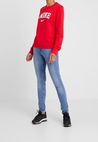 Röd sweatshirt med vitt Nike-logotyp, rund halsringning och ribbad mudd och nederkant. Kombineras med ljusblå jeans och svarta löparskor.