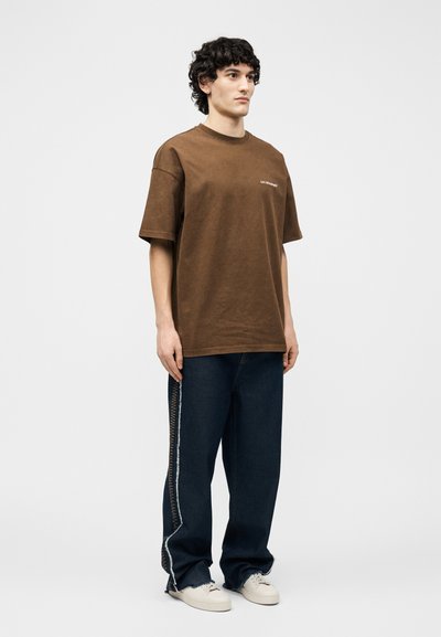 Νέος άνδρας στέκεται μπροστά, φοράει άνετο καφέ t-shirt, σκούρο τζιν με πλαϊνές ραφές και λευκά αθλητικά παπούτσια σε απλό φόντο.