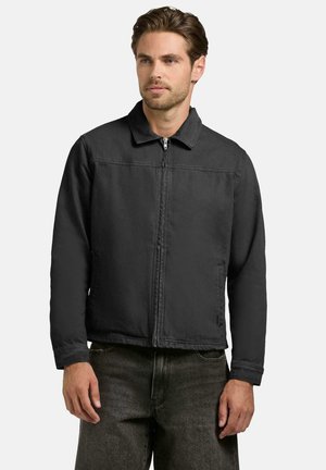 Mężczyzna w czarnej kurtce zapinanej na zamek z kołnierzem i bocznymi kieszeniami, ubrany w ciemnoszare spodnie, stoi na jednolitym jasnym tle.