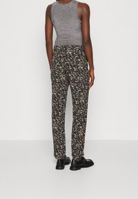 Grijze mouwloze top en zwarte broek met een patroon in crèmekleuren en bruintinten. Gladde textuur, elastische tailleband en rechte snit. Zwarte schoenen.
