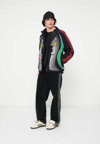 Ung mann iført svart caps, grafisk zip-up jakke med røde striper, svarte bukser med sidestriper og hvite Adidas sneakers, står mot en hvit vegg.