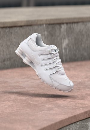 Balta sportinė sneaker su išskirtiniu paminkštintu padu, sklandanti virš rausvo betono paviršiaus ir pilko akmens fono.