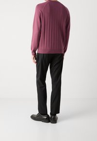 Un homme portant un pull violet à col rond à manches longues, un pantalon noir et des chaussures de costume noires, se tenant sur un fond blanc uni.