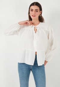 Bijela bluzica dugih rukava s vezicom oko vrata, kopčanje na gumbe, nabrani yoke i peplum rub, nosi se s plavim trapericama. Mekana, strukturirana tkanina.
