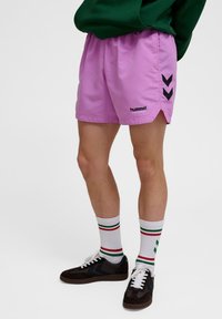Ljusrosa shorts med dragsko i midjan, med svart "hummel"-logotyp och chevron-accenter. Kommer med vita strumpor och mörka skor.