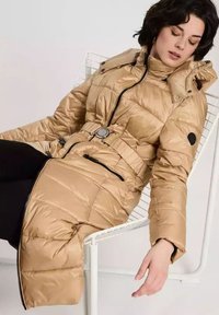 Puchowa złota parka z paskiem w talii, z zamkiem błyskawicznym i kapturem. Posiada pikowaną teksturę i czarny okrągły akcent na lewym rękawie.