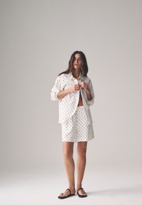 Weißer Schlafanzug mit schwarzen Punkten. Besteht aus einem langärmligen Hemd mit Knopfleiste und passenden Shorts. Glatter Stoff, lockere Passform.