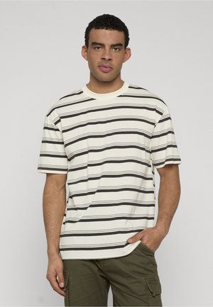 Homme aux cheveux courts et bouclés portant un t-shirt à rayures horizontales blanches et noires et un pantalon cargo vert olive, une main dans la poche.