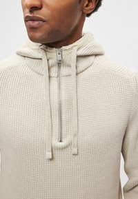 Sweatshirt bege claro com capuz, com padrão texturizado em malha canelada, apresentando fecho de correr frontal e capuz com cordão. Acabamento em tecido macio.