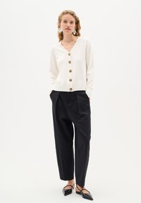 Elfenbenfärgad stickad cardigan med v-ringning, ribbad fåll och guldknappar; kombinerad med svarta högmidjade byxor och svarta spetsiga ballerinas.