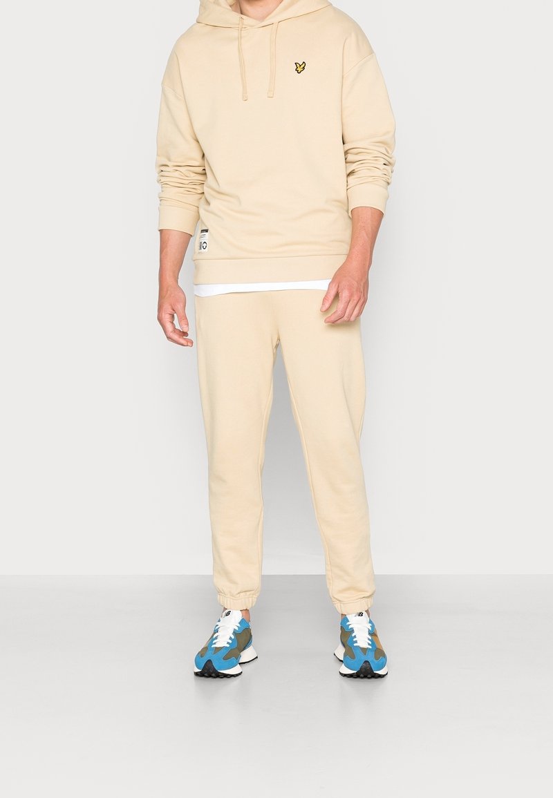 Homme portant un hoodie beige et un pantalon de survêtement assorti avec des baskets bleues, vertes et blanches, debout devant un fond uni.