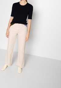 Marineblaues, kurzes Strickoberteil mit kontrastierenden Manschettendetails, kombiniert mit hellbeigen, weit geschnittenen Hosen und cremefarbenen Schuhen mit quadratischer Spitze auf hellem Hintergrund.