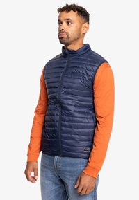 Chaleco acolchado azul marino con acolchado horizontal, cuello alto y cremallera frontal; usado sobre una camiseta de manga larga naranja. Presenta un detalle de logo en el dobladillo.