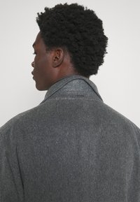 Cappotto di lana grigia con una consistenza morbida, caratterizzato da un colletto ampio e un discreto logo del brand sul retro. Il design è semplice e moderno.