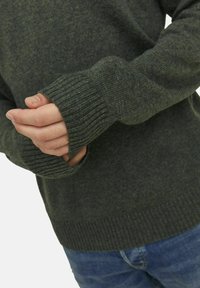 Dunkelgrüner gestrickter Pullover mit gerippten Bündchen und Saum, der eine weiche Textur und mittlere Dicke aufweist. Die Hände sind in der Nähe der Taille platziert.