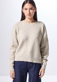 Beige gebreide trui met een diamantpatroon, ronde halslijn en geribbelde manchetten. G gedragen met donkerblauwe wijde broek. Zachte textuur.