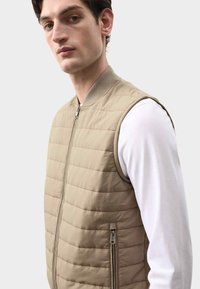 Chaleco acolchado beige con cremallera frontal, cuello acanalado y costuras horizontales. Se lleva sobre una camisa blanca de manga larga y cuenta con bolsillos laterales.