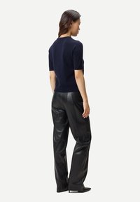 Kvinna vänd mot bort, klädd i marinblå kortärmad tröja, svarta vida byxor i läder och svarta spetsiga klackar mot en vit bakgrund.