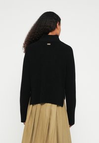 Fekete kötött pulóver magas gallérral, bordázott mintázattal és arany logóval a háton. Rövidített hosszúságú, hosszú ujjakkal.